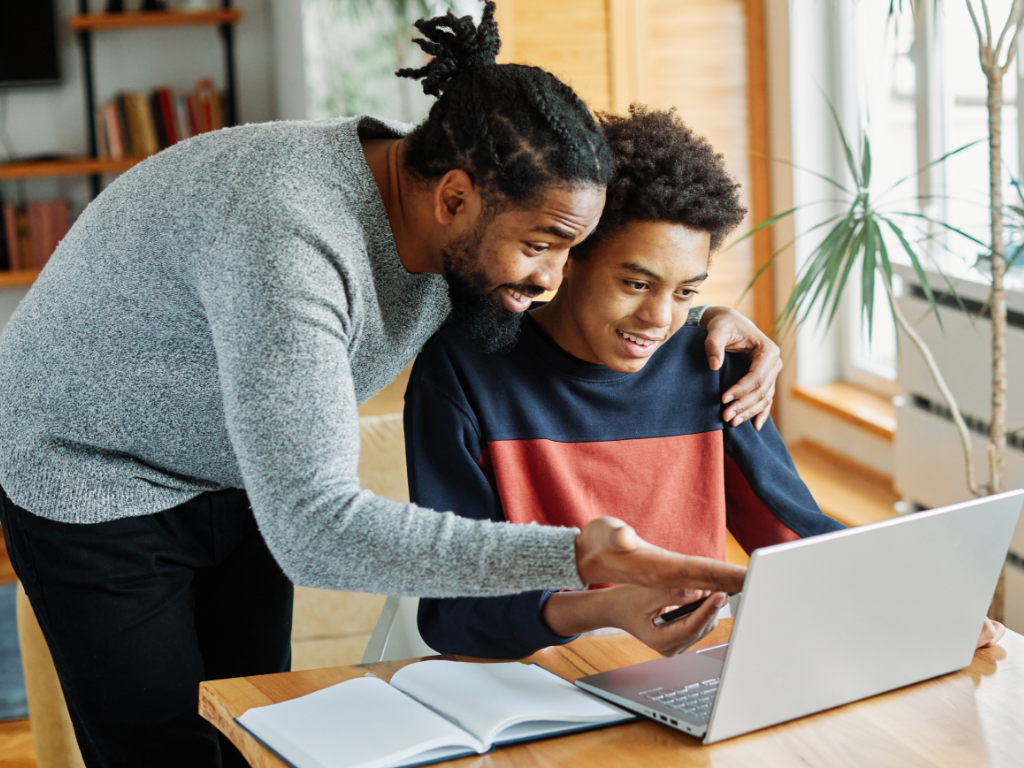 A teen boy sits at a table with a pen and notebook, looking at a computer, while his dad looks over his shoulder pointing at something on the screen to signify parents looking for opportunities to teach their children how about investing